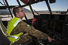 Bespectacled man in camouflage uniform with fluorescent jacket in cockpit of military aircraft