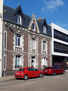 Cliché numérique couleur. Devant une maison de la fin du XIXe siècle et un immeuble du XXe siècle de trois étages stationnent deux voitures rouges.