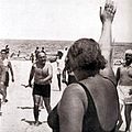 Femme de dos, en costume de bain, faisant le salut fasciste au Duce (en short noir) sur la plage de Riccione où sont présent quelques autres personnages en vêtements de plage.