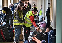 Two young people in hi-viz jackets hand out bottled drinks as other people pass by with luggage