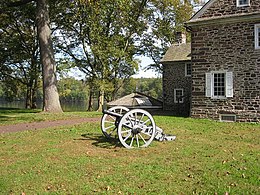 Photo shows an 18th-century cannon, probably a 3-pounder, with an old stone building and the Delaware River in the background.