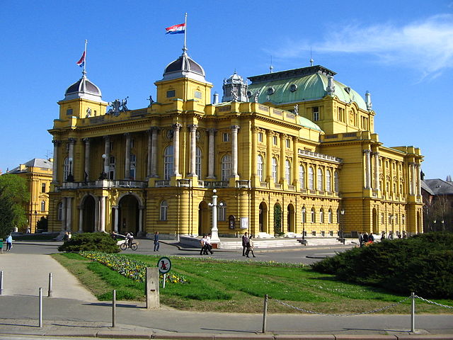 Żółty duży budynek na placu, nad nim powiewa flaga Chorwacji. Jest piękny słoneczny dzień.