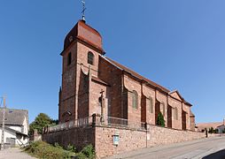 Igreja de arenito rosa de três quartos.  Sucessivamente: campanário Comtois em azulejos vermelhos, ábside sem transepto.