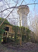 Большая бетонная водонапорная башня выступает из разрушенного производственного здания из кирпича и бетона в лесу.