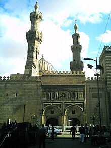 Duas entradas em arco no pórtico de um grande edifício de dois andares voltado para uma rua.  Acima dos arcos, a parede do edifício é entalhada e ornamentada.  À direita, o edifício sobe para um terceiro andar.  Atrás da parede, dois minaretes emoldurando o topo de uma cúpula são visíveis.