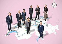 The Swiss Federal Council in 2022 with President Ignazio Cassis (bottom) standing on an abstract, reduced railway lines map and positioned at their respective political origins Bundesratsfoto 2022.jpg