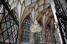 vue en contre plongée et à travers une grille entrouverte de l’intérieur d’une chapelle, avec quatre grandes fenêtres dotées de vitraux et une voûte réticulée.