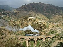 Ligne de chemin de fer érythréenne avec une locomotive à vapeur noire passant sur un pont en pierre.