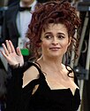 A woman with long, curly auburn hair in a black dress, waving