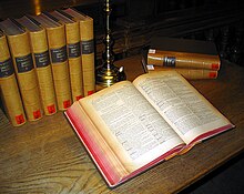 Photograph of an open volume of a dictionary lying on a table, with other volumes standing or lying around it.