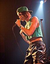 An image of a woman performing on a stage. She is wearing a crop top, shiny shorts, and a baseball cap. She is singing into a microphone and looking away from the camera.