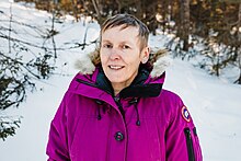 Photo of Lela Evans, NDP MHA for the District of Torngat Mountains, wearing a magenta coloured Canada Goose Parka with snow and spruce trees behind