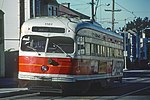 Streetcar No. 1101 of the San Francisco Municipal Railway in 1980. The streetcar is painted in the "Sunset" livery designed by Walter Landor, which is primarily white with a reddish orange horizontal stripe encircling the streetcar below the window line, itself bisected by a thin yellow-orange stripe.