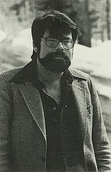 Un homme brun barbu avec de grosses lunettes.
