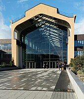 Photo de l'entrée principale de la Cité du Cinéma. On voit un bâtiment de type industriel rénové et possédant une façade en verre située devant un parvis.
