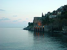 Uma colina povoada de casas desce em um mar azul esverdeado abaixo de um cais de pedra com cinco arcos.  Uma parede de pedra se estende ao longo do mar, desde o cais até o canto inferior direito.