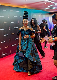 A woman with medium-dark skin and towering coils of black hair stands regally on a red carpet in a flowing, off-the-shoulder blue and black gown.
