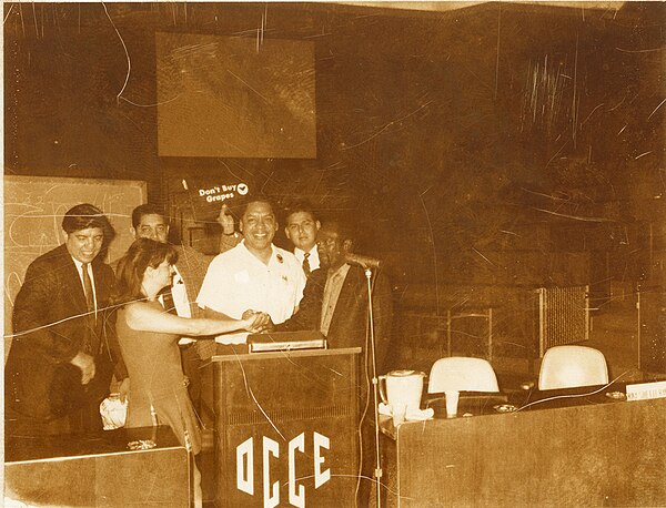 Francisco 'Pancho' Medrano speaks at conference on the boycott of grapes around 1965–1967. In the back, an unidentified man holds a sign that reads "D