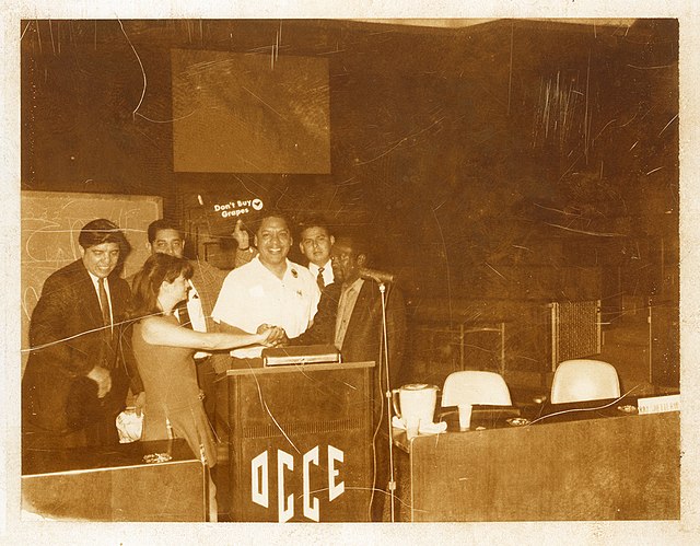 Francisco 'Pancho' Medrano speaks at conference on the boycott of grapes around 1965–1967. In the back, an unidentified man holds a sign that reads "D