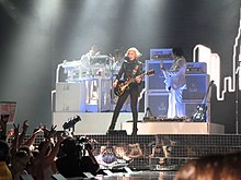 Sarışın bir kadın sanatçı sahnede elektrikli gitar çalarken. Siyah bir ceket ve pantolon giymiş. Arkasındaki görüntülerde yüksek binalar gösteriliyor. Sahne, bir dizi halojen lamba ile aydınlatılmış.