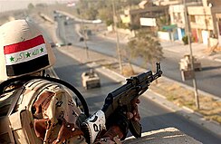 An Iraqi Army officer, whose helmet bears a painted flag of Iraq, holds a rifle as he overlooks a market across a road. There are two tanks placed on the road in question.