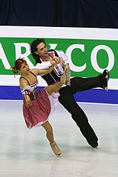 Un homme et une femme dansant sur glace ;  la femme, à gauche, a les cheveux roux tirés en arrière et porte une robe paysanne rose foncé et blanche, et l'homme porte une chemise noire et blanche avec des revers rose foncé et un pantalon noir