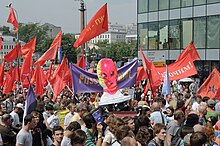 Amid a sea of protesters, someone holds an oversize Styrofoam head clad in a multicolored ski mask. Behind it is a sign saying 