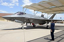 Foto a colori di un aereo militare grigio a terra.  Un uomo vestito di blu è in piedi vicino e saluta l'aereo.