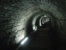 A dimly-lit brick-lined tunnel about 2m tall and slightly less wide, curving away gradually to the right, with lights at intervals along the wall.