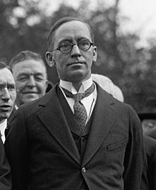 Black and white photograph of man with short, wispy hair wearing circular glasses, necktie, suit, and overcoat