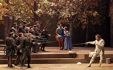 Theatre scene with men in 15th-century costume facing off with drawn swords, while a young couple embrace protectively at the rear of the stage