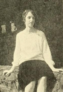 A young white woman, seated outdoors, wearing a white blouse and a dark skirt