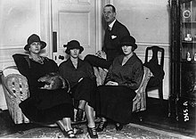 Photographie en noir et blanc montrant un homme assis derrière un canapé sur lequel sont assises trois femmes portant chapeaux.