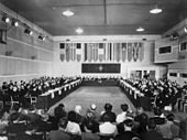 Unha longa sala rectangular con varias filas de individuos sentados a cada lado, e bandeiras colgadas no extremo.