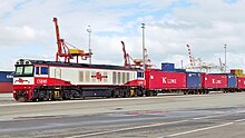 Red and white diesel locomotive hauling intermodal freight wagons in a shipping port with cranes behind