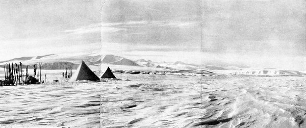 Panoramic photograph of a camp with two tents and a line of skis on ice with a sprawling mountain in the distance