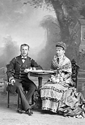 Photographie en noir et blanc d'un couple pose assis près d'un guéridon, l'homme est habillé en officier et sourit, une cigarette à la main, la dame porte une ample robe se tient les mains croisées, sans regarder l'objectif du photographe.