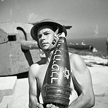 Joseph Wald, a Jewish Brigade soldier, carries an artillery shell. The Hebrew inscription on the shell translates as "A gift to Hitler." Jewish Brigade-gift.jpg