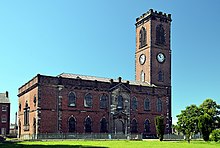 A two-storey brick church with stone dressings seen from the north; the tall slim tower has clock faces and a battlemented parapet