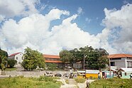 In the foreground there is a large building with red roofs. There are trees surrounding the it. The sky is blue with some white clouds.