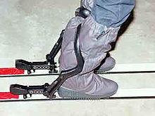 A pair of Nava System bindings with the skier in place. The boot is clipped into the binding, which is almost flush with the ski, and the control arm, black, is in place behind the skiers calf. Nava bindings.jpg