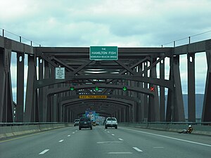 A roadway with large brown metal latticework above it. On the side facing the camera is a small white on green sign reading "The Hamilton Fish Newburgh-Beacon Bridge"