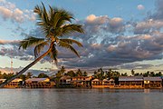 River bank of the island of Don Khon with stilt wooden houses, seen from Don Det with a leaning Arecaceae (palm trees) and colorful clouds