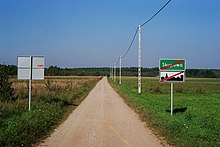 Przez łąkę prowadzi prosta droga o nawierzchni piaszczystej lub żwirowej. Wzdłuż niej ustawiona jest linia telefoniczna lub energetyczna. W oddali widoczny las.