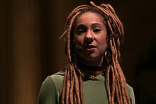 Mulher negra, com dreads compridos. Possui um piercing de argola no nariz, usa brincos de argola e está vestindo uma blusa sem estampa de cor verde musgo