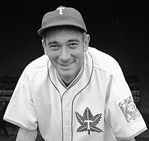 Un hombre, con una gorra de béisbol con una "T" en el centro y un uniforme de béisbol blanco con el logo de béisbol de los Toronto Maple Leafs en el pecho izquierdo, se inclina hacia adelante sonriendo.