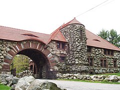 The exterior of this gate house is made of rough stone and finished brownstone. It has a tile roof, in which two low eye windows are built. A road lined with rough stone walls passes through an arch in the structure.