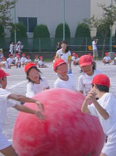 Crianças vestidas de branco e com bonés vermelhos empurram uma grande bola vermelha.