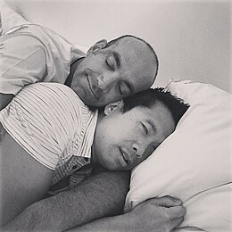 Black and white photograph of two men (clothed) cuddling in bed.jpg