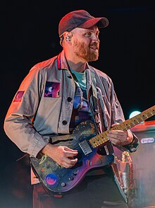 A man wearing a dark blue cap and jacket playing a guitar